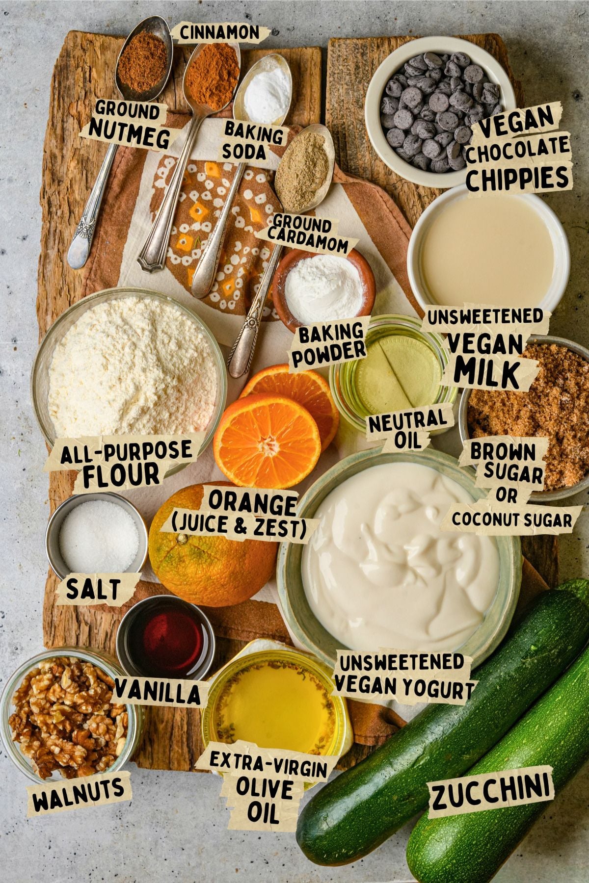 Ingredients for a vegan zucchini muffin recipe including zucchini, flour, spices, orange, vegan yogurt, vegan milk, walnuts, vanilla, olive oil, sugar, baking powder, and chocolate chips, are arranged on a wooden board.
