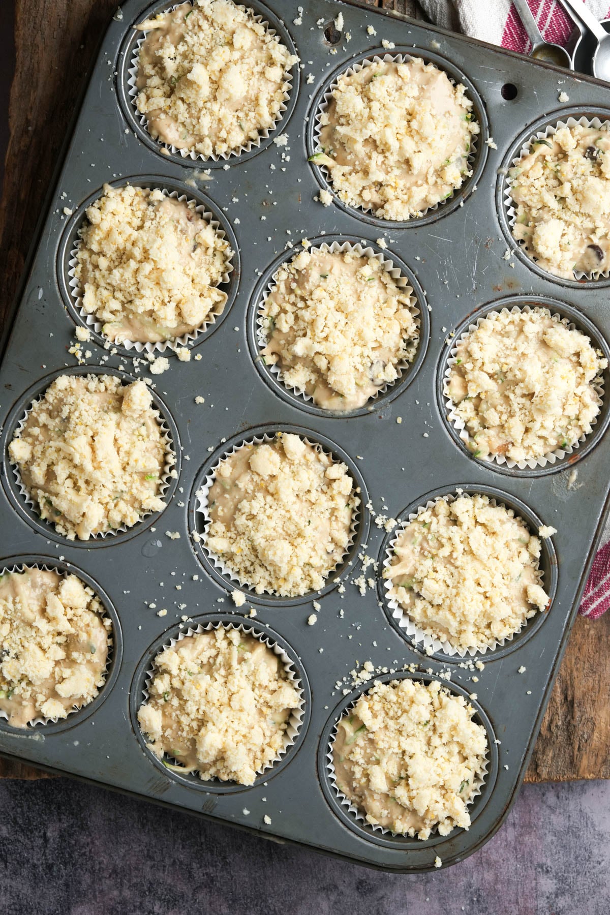 A metal muffin tin holds twelve unbaked vegan zucchini muffins with crumbly streusel topping in paper liners, ready to go into the oven. The tin rests on a wooden surface near a red-striped kitchen towel.