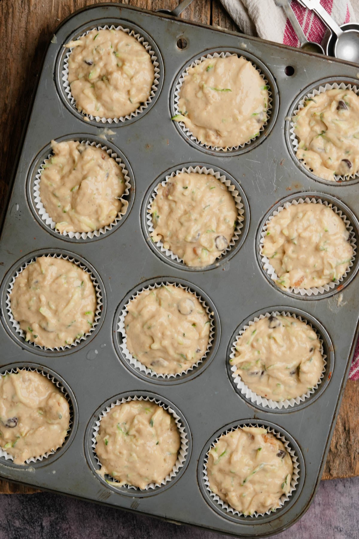 A muffin tin filled with paper liners, each holding unbaked vegan zucchini muffin batter with visible bits of zucchini and nuts, sits on a rustic wooden surface.