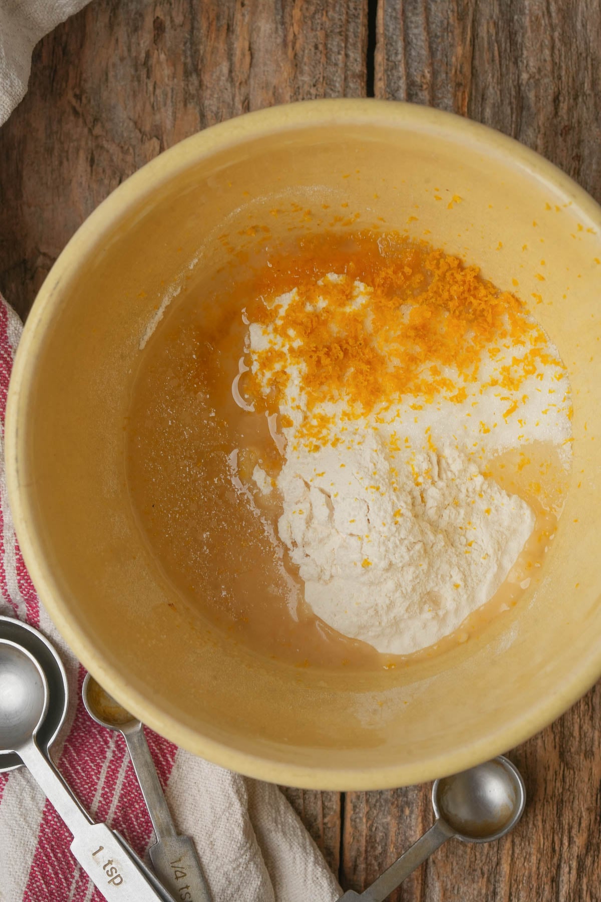 A yellow mixing bowl with flour, liquid, and orange zest inside sits on a rustic wooden surface beside metal measuring spoons and a red-striped cloth.