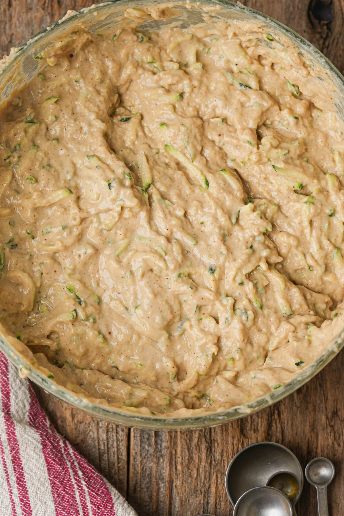 Bowl of thick vegan zucchini muffin batter with visible shredded zucchini, on a rustic wooden surface next to a red-striped dish towel and measuring spoons.