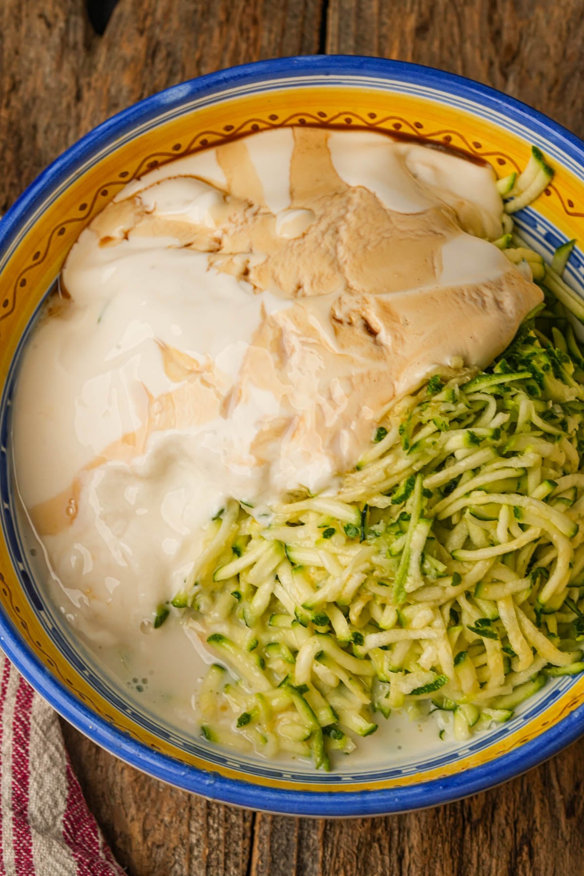 A colorful bowl with shredded zucchini, vegan yogurt, vegan milk, oil, and vanilla, all unmixed, sits on a rustic wooden surface next to a red-striped cloth.