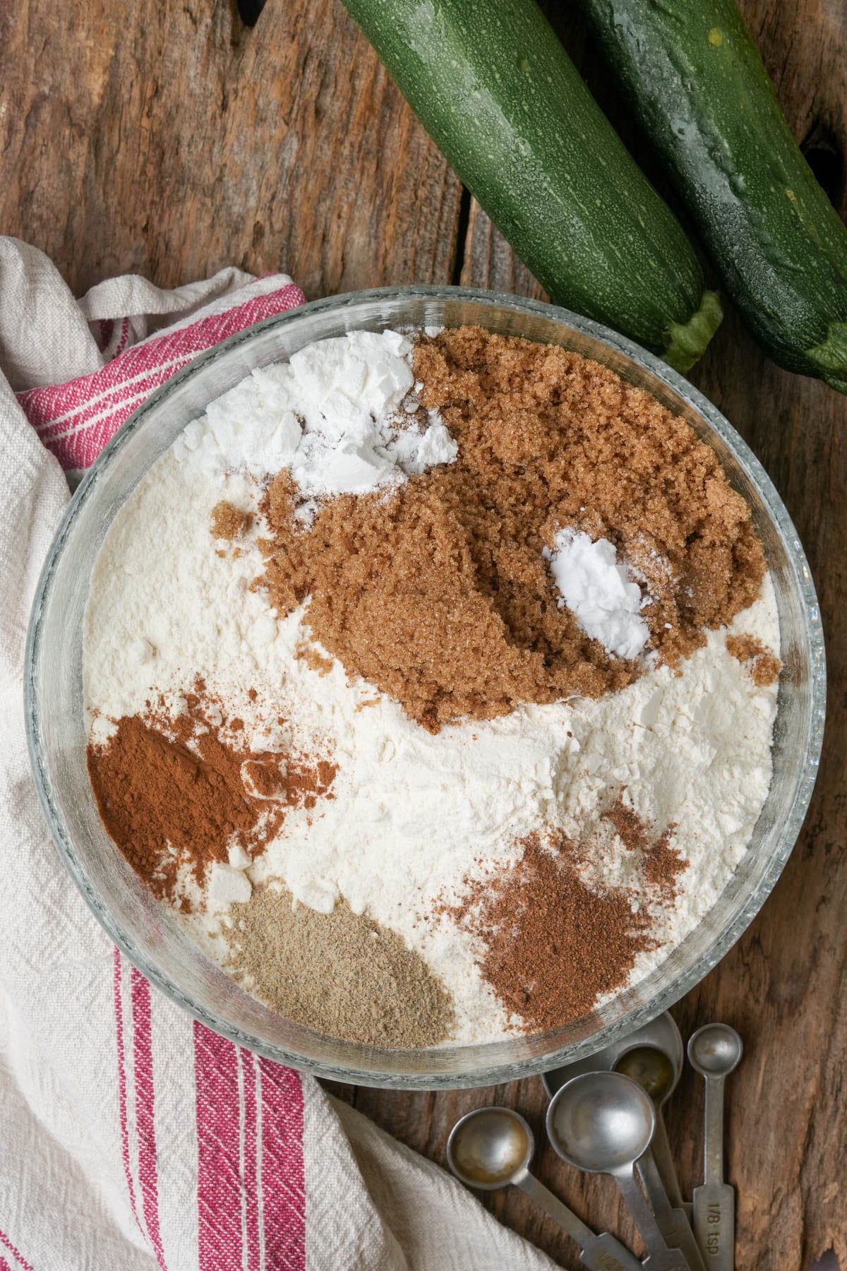 A bowl with flour, brown sugar, spices, and leavening agents on a wooden table, next to two zucchinis, measuring spoons, and a striped white towel.