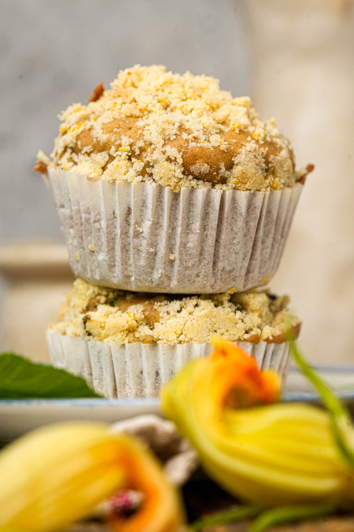 Two crumb-topped vegan zucchini muffins stacked on top of each other, wrapped in white paper liners, with a blurred yellow flower and green leaf in the foreground.