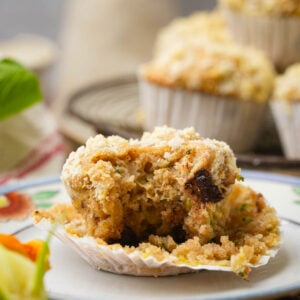 A partially eaten vegan zucchini muffin with crumbs sits on a plate, with more whole muffins in the background on a cooling rack. The muffin appears moist with pieces of fruit or nuts visible.