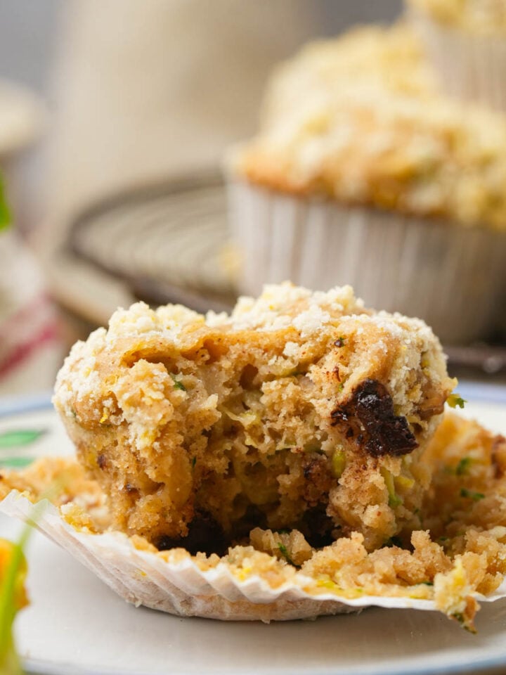 A partially eaten vegan zucchini muffin with crumbs sits on a plate, with more whole muffins in the background on a cooling rack. The muffin appears moist with pieces of fruit or nuts visible.