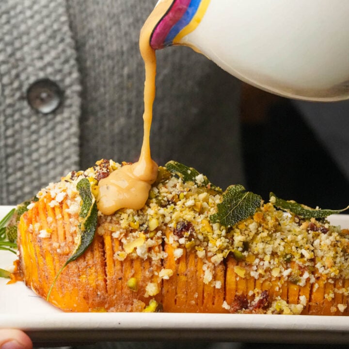 A person pours sauce over a sliced, baked Hasselback butternut squash topped with breadcrumbs, nuts, and crispy sage, served on a white dish.