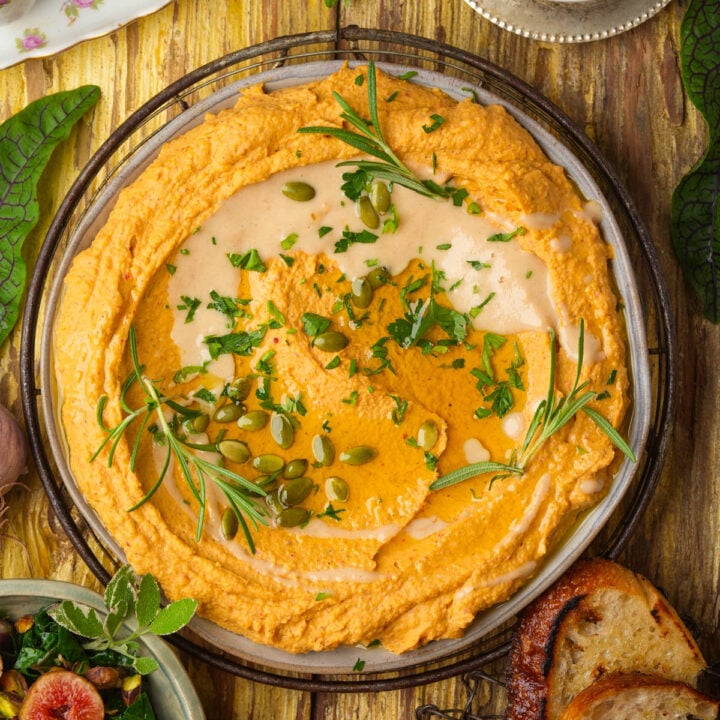 A bowl of creamy butternut squash hummus garnished with herbs, pumpkin seeds, and olive oil, surrounded by toasted bread slices and salad on a rustic wooden table.