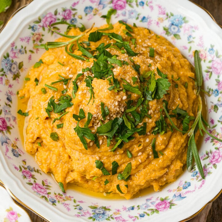 A bowl of creamy orange mashed butternut squash topped with chopped parsley, sesame seeds, and herbs, served in a floral-patterned dish.