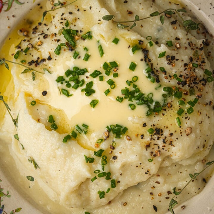 Creamy mashed cauliflower topped with melted butter, chopped chives, cracked black pepper, and fresh thyme, served in a bowl.