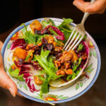 A hand holds a colorful roasted pumpkin salad with leafy greens, baked pumpkin, walnuts, dried cranberries, and fresh herbs on a patterned plate, with a fork ready to eat.