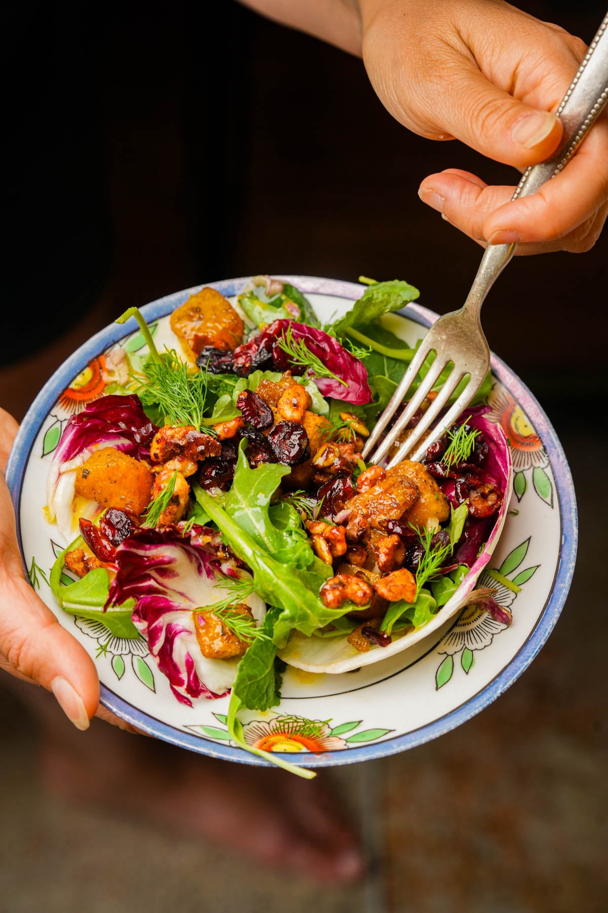 A person holds a decorative bowl of roasted pumpkin salad with greens, roasted pumpkin, walnuts, cranberries, and fresh herbs, using a fork to take a bite.