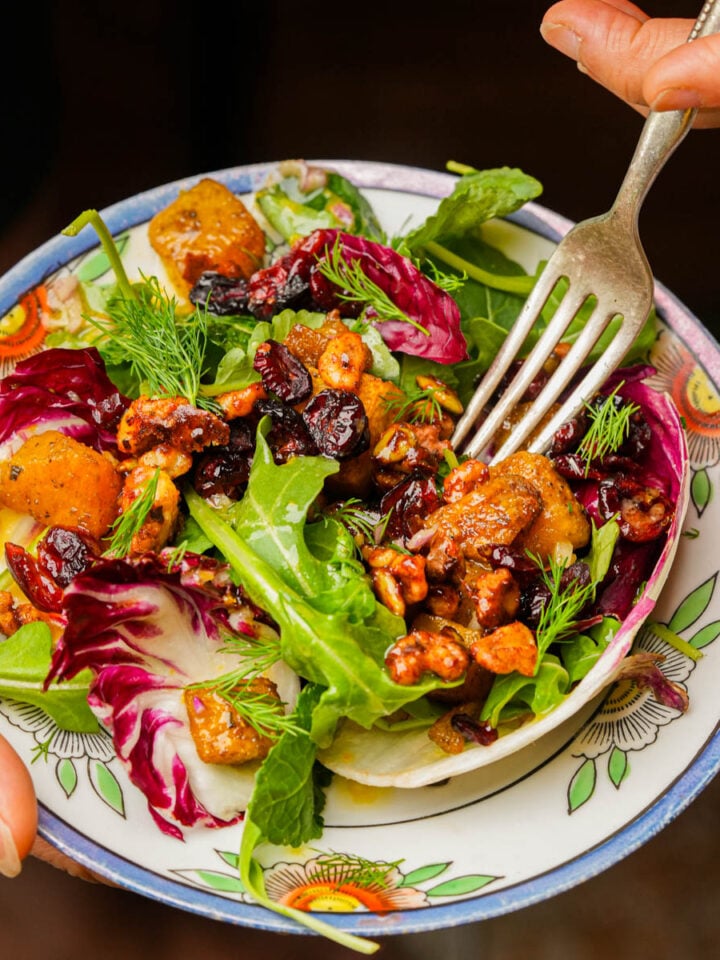 A hand holds a colorful roasted pumpkin salad with leafy greens, baked pumpkin, walnuts, dried cranberries, and fresh herbs on a patterned plate, with a fork ready to eat.