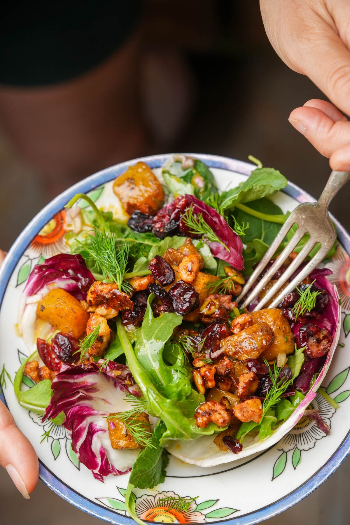 A colorful roasted pumpkin salad with mixed greens, roasted pumpkin, candied nuts, dried cranberries, and fresh dill, served on a decorative plate, with a hand holding a fork ready to take a bite.