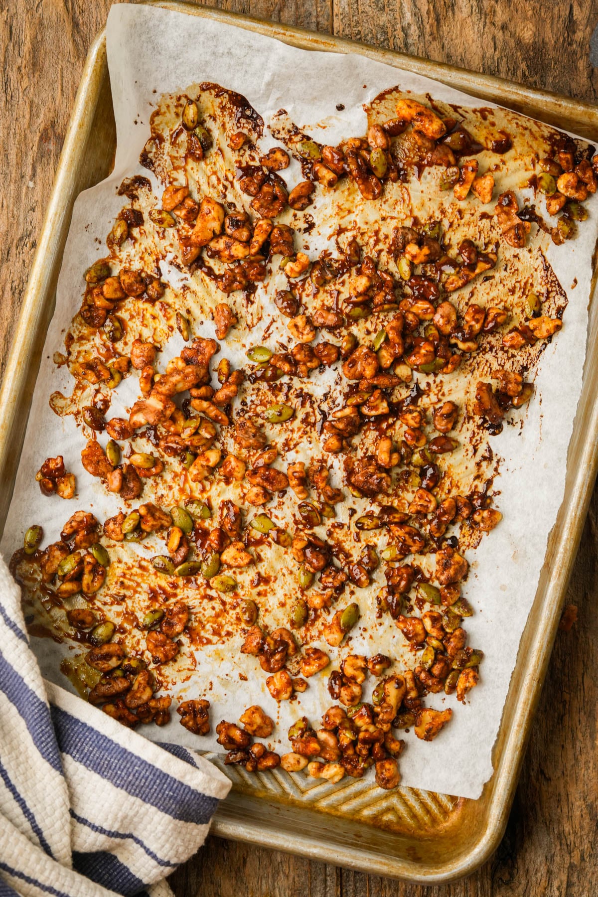 A baking sheet with parchment paper holds roasted nut and seed clusters coated in spices. A striped towel is on the corner of the tray, all set on a rustic wooden surface.
