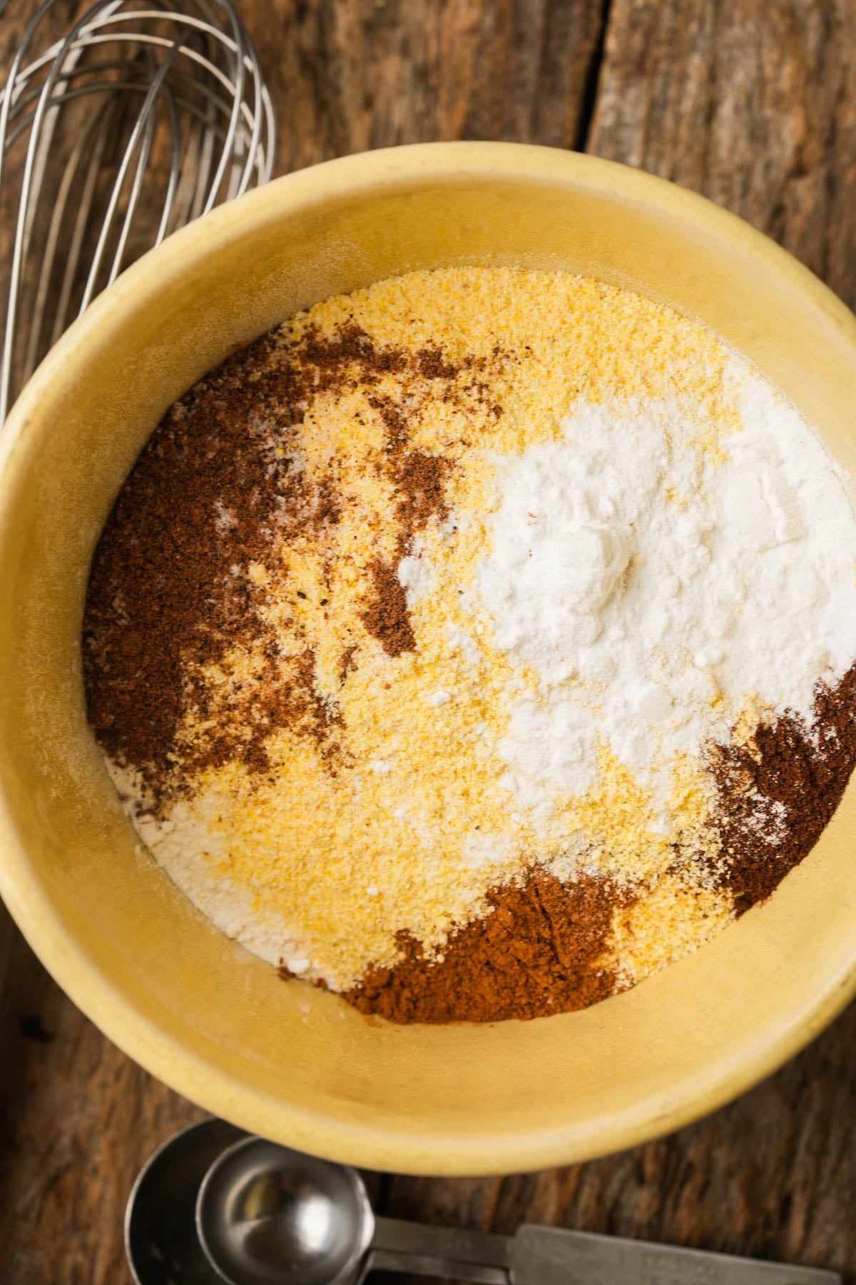 A yellow bowl on a wooden surface holds dry ingredients for a vegan pumpkin cornbread recipe. A metal whisk and measuring spoons are nearby.