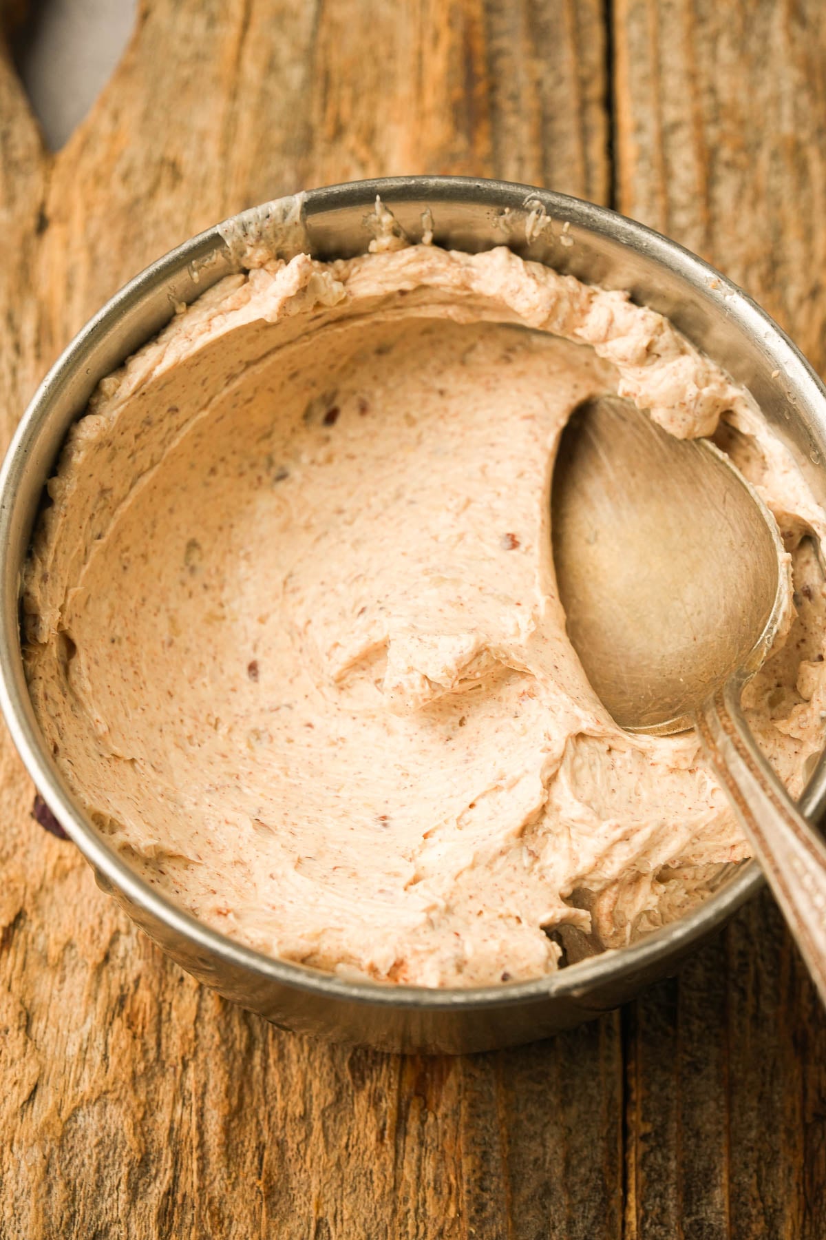 A metal bowl filled with cinnamon pecan butter with a spoon resting inside, placed on a rustic wooden surface.
