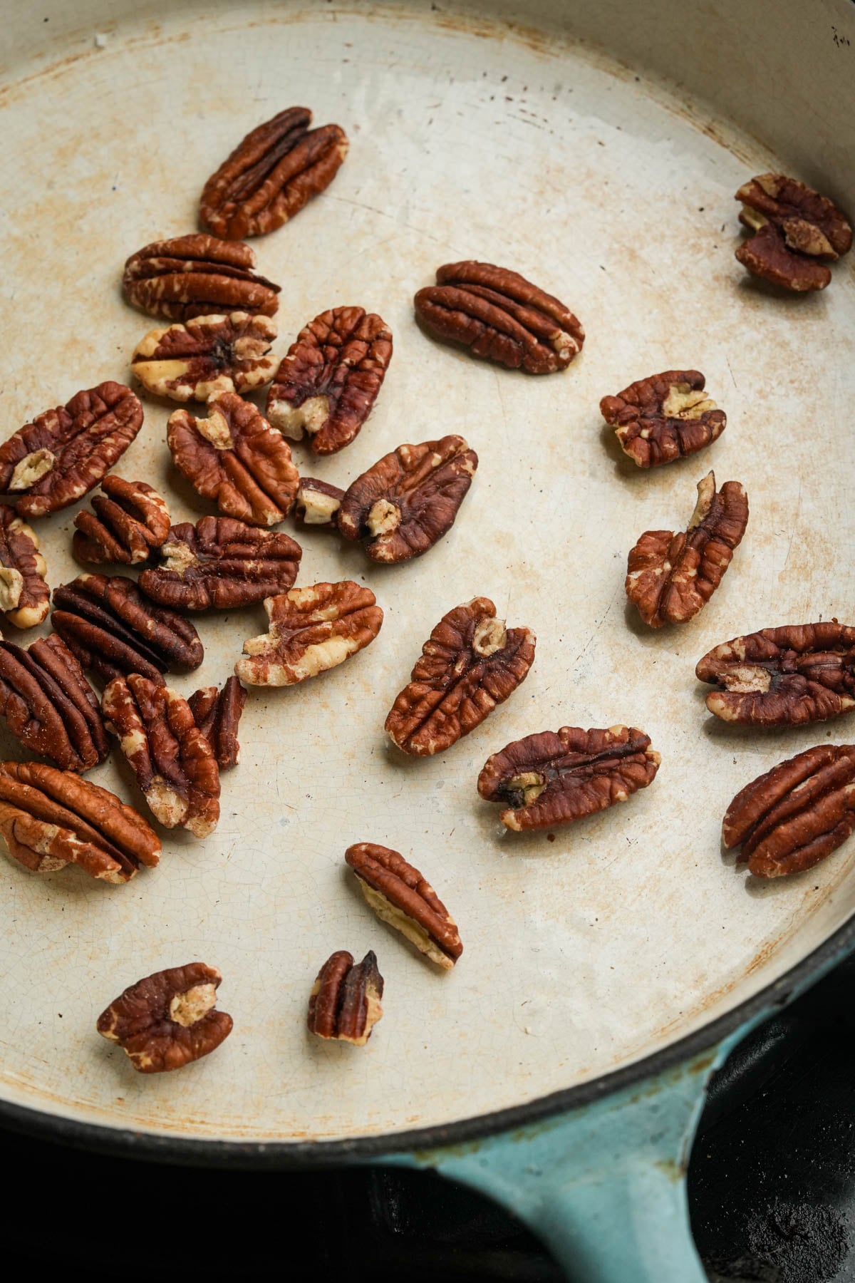 Pecan halves scattered in a light-colored skillet, some toasted, with a turquoise handle visible at the bottom right corner.