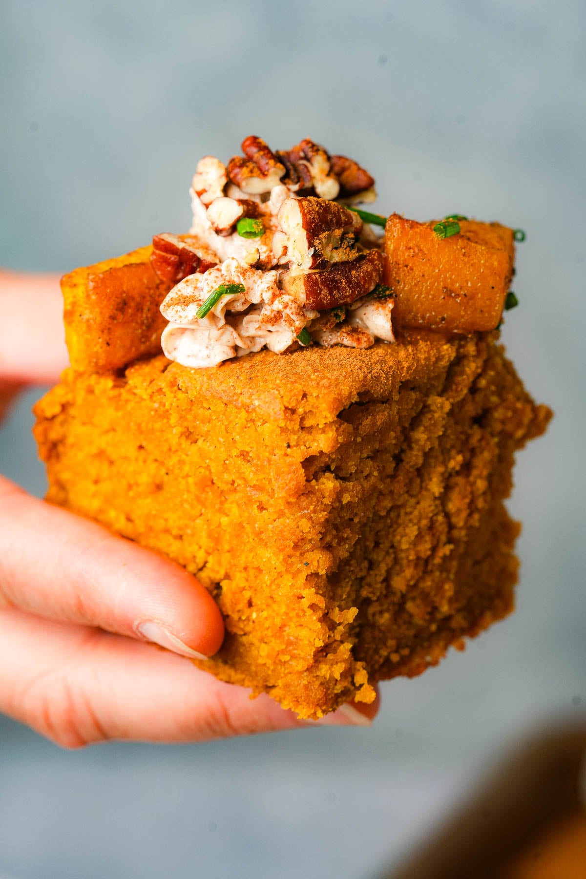 A hand holds a thick slice of vegan pumpkin cornbread topped with whipped cinnamon pecan butter, roasted pumpkin cubes, chopped pecans, and herbs against a blurred background.