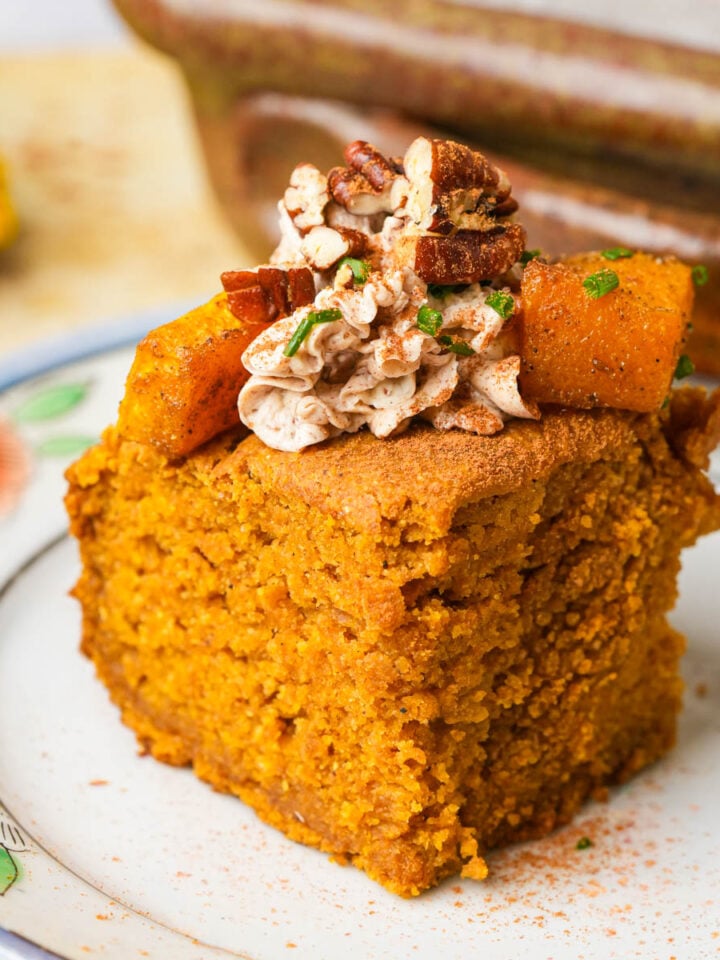 A slice of vegan pumpkin cornbread topped with spiced whipped butter, pecans, and pumpkin cubes sits on a decorative plate.