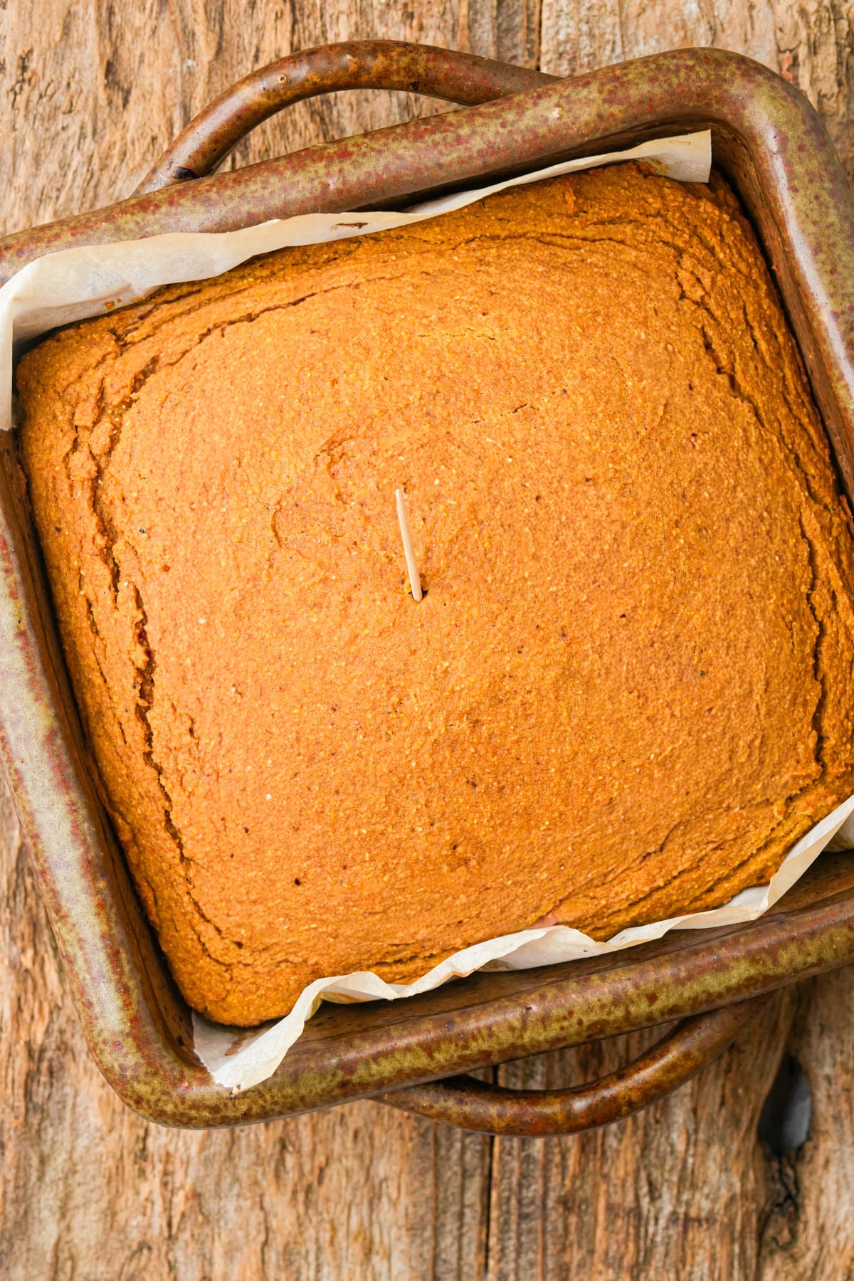 A freshly baked square vegan pumpkin cornbread in a ceramic pan with parchment paper, shown from above. A toothpick is inserted in the center to check doneness. The cake has a golden-brown, slightly cracked top and sits on a rustic wooden surface.