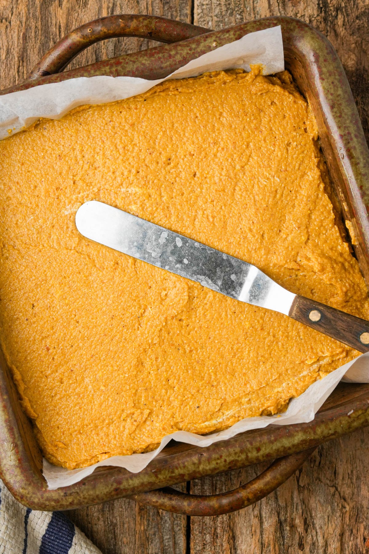A baking dish lined with parchment paper holds a smooth layer of vegan pumpkin cornbread batter. A metal spatula with a wooden handle rests on top of the batter. The dish sits on a rustic wooden surface.