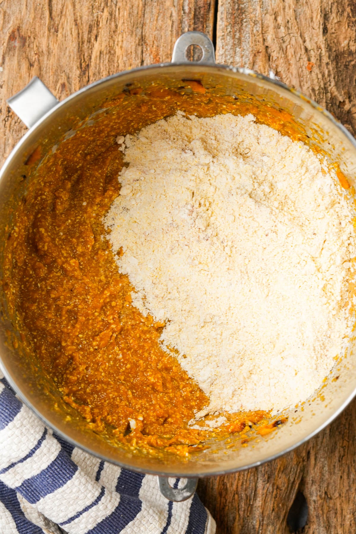 A metal mixing bowl with orange vegan pumpkin cornbread batter with dry ingredients partially mixed in, sitting on a rustic wooden surface next to a blue and white striped cloth.