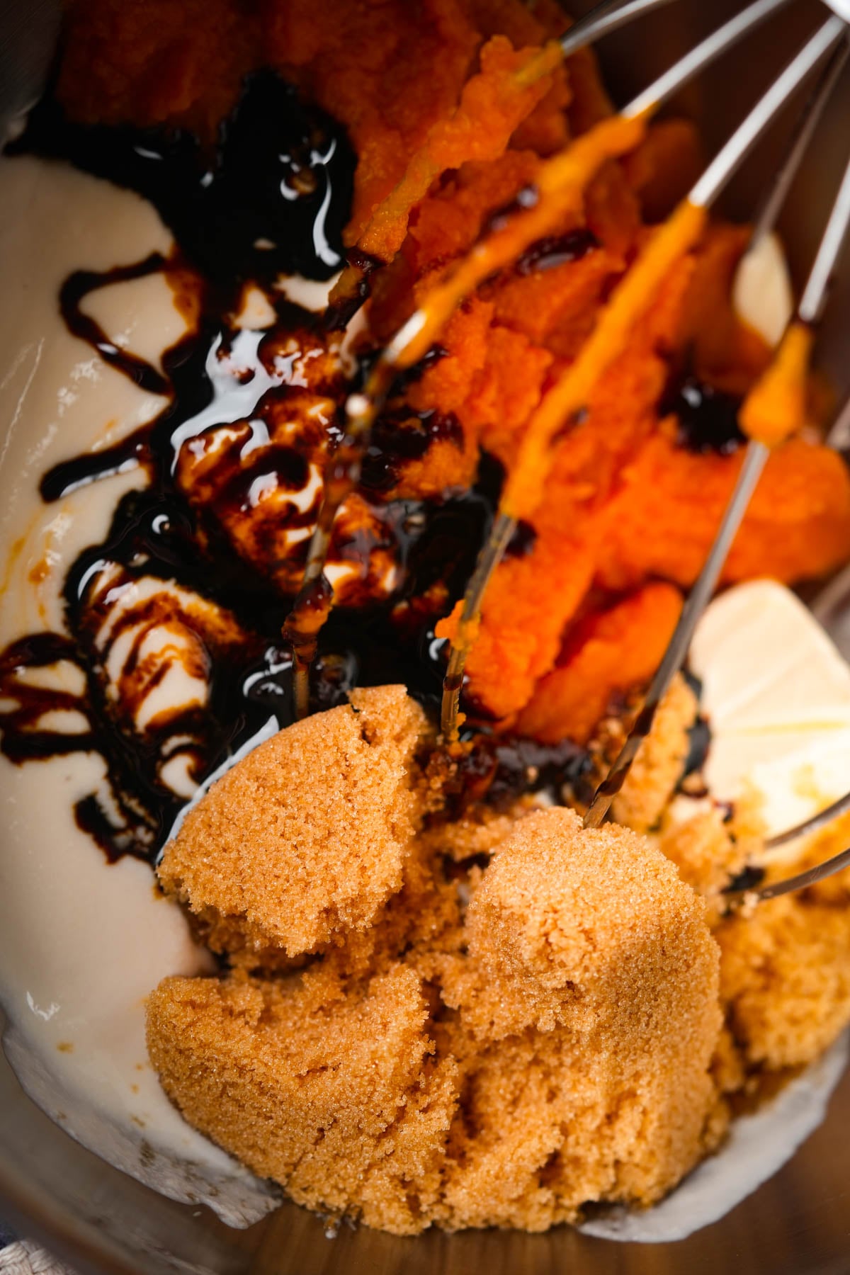 Close-up of a mixing bowl containing brown sugar, pumpkin puree, dark molasses, and vegan yogurt, with a whisk partially blending the mixture.