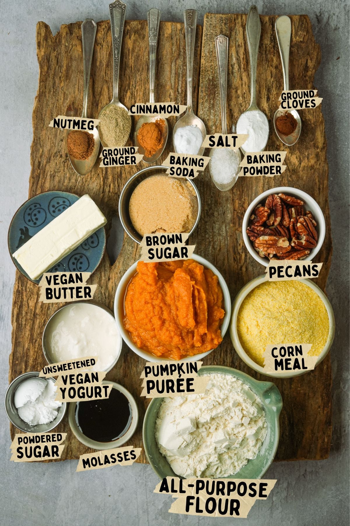A wooden board holds labeled spoons of spices and bowls of vegan butter, yogurt, brown sugar, pumpkin purée, pecans, cornmeal, flour, powdered sugar, and molasses, all ingredients for a vegan pumpkin cornbread recipe.