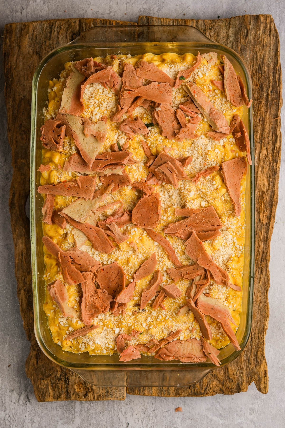A glass baking dish filled with unbaked vegan pumpkin macaroni and cheese, topped with breadcrumbs and slices of vegan bacon, sits on a wooden board.
