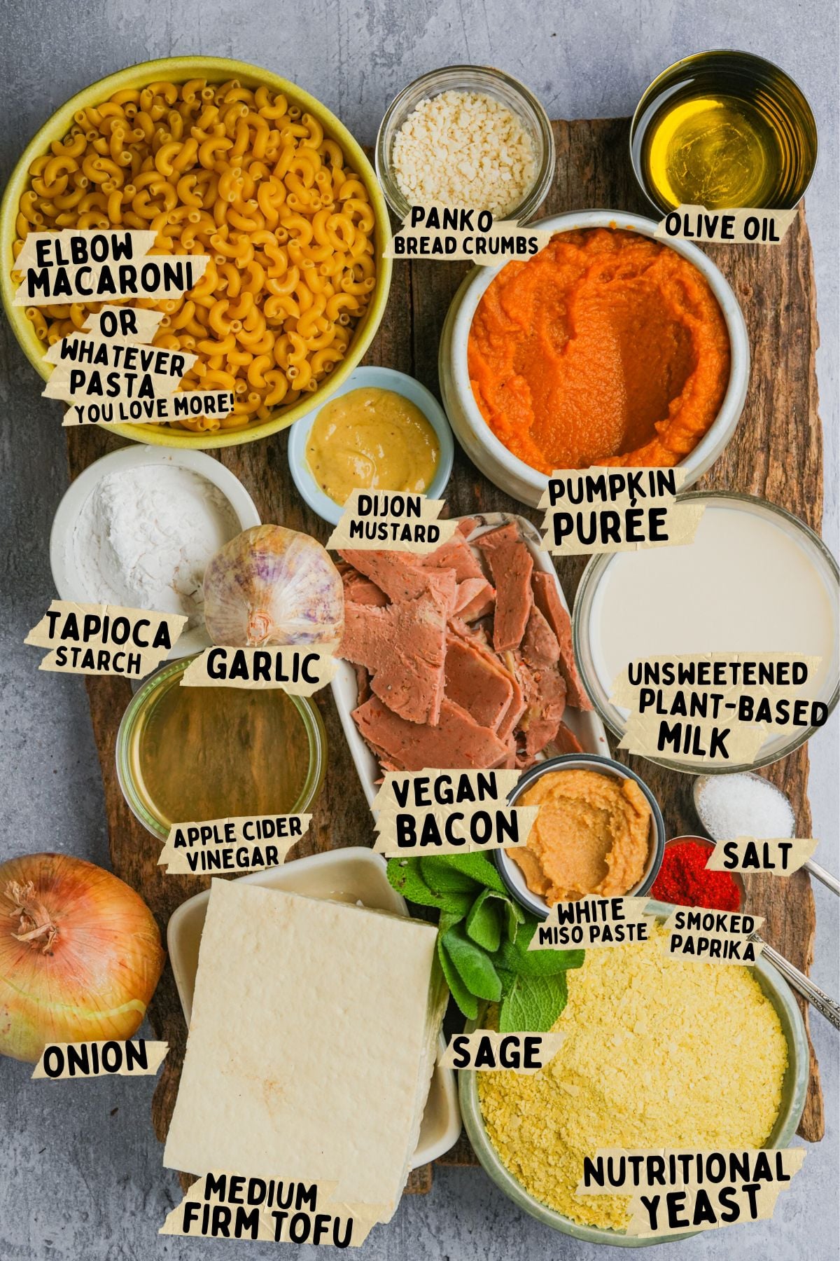 A variety of ingredients on a wooden board, including elbow macaroni, tofu, plant-based milk, nutritional yeast, vegan bacon, pumpkin puree, spices, onion, garlic, and oil, all labeled for making vegan pumpkin mac and cheese.