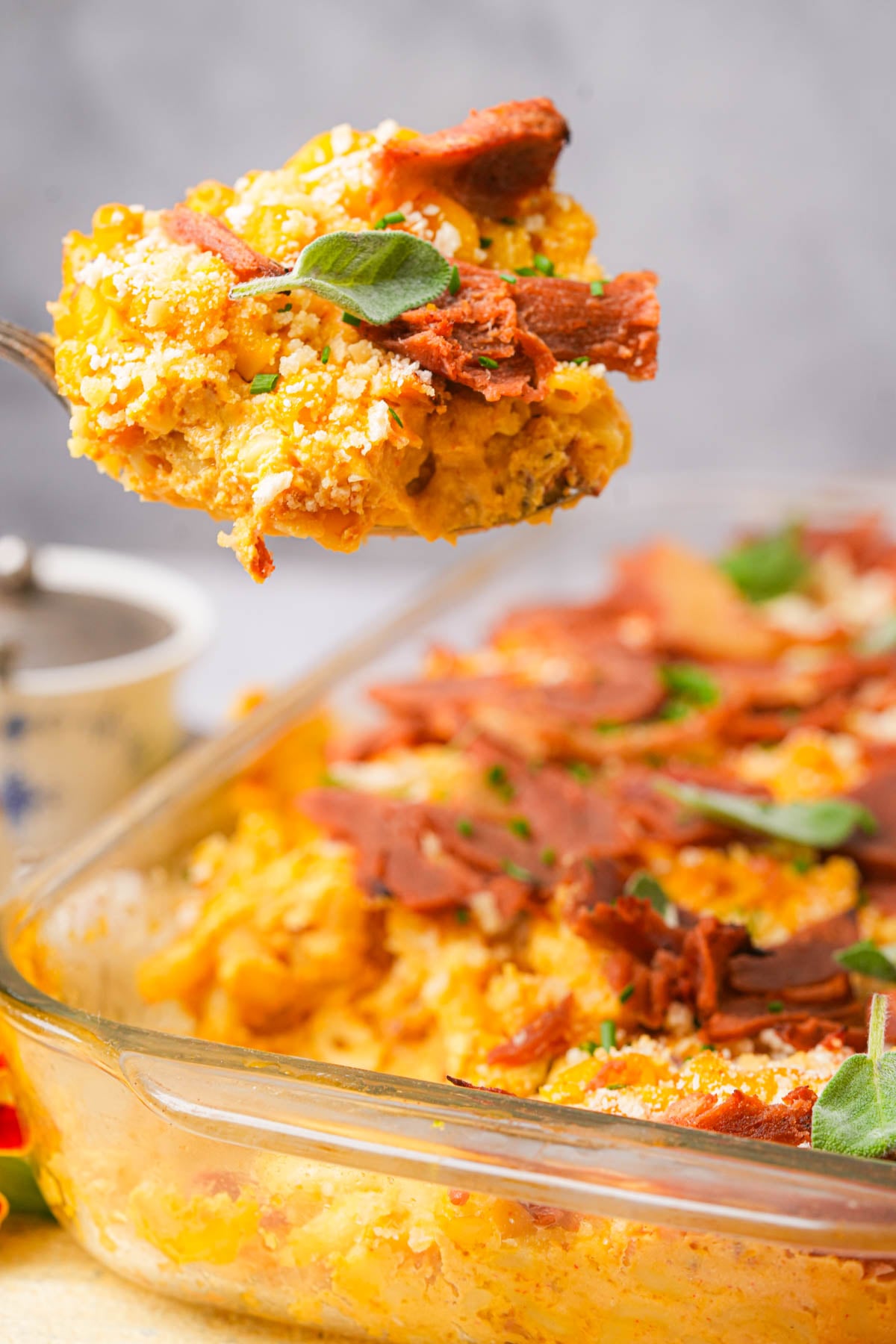 A spoon lifting a portion of cheesy vegan pumpkin mac and cheese topped with crispy bacon from a glass baking dish, garnished with fresh herbs.