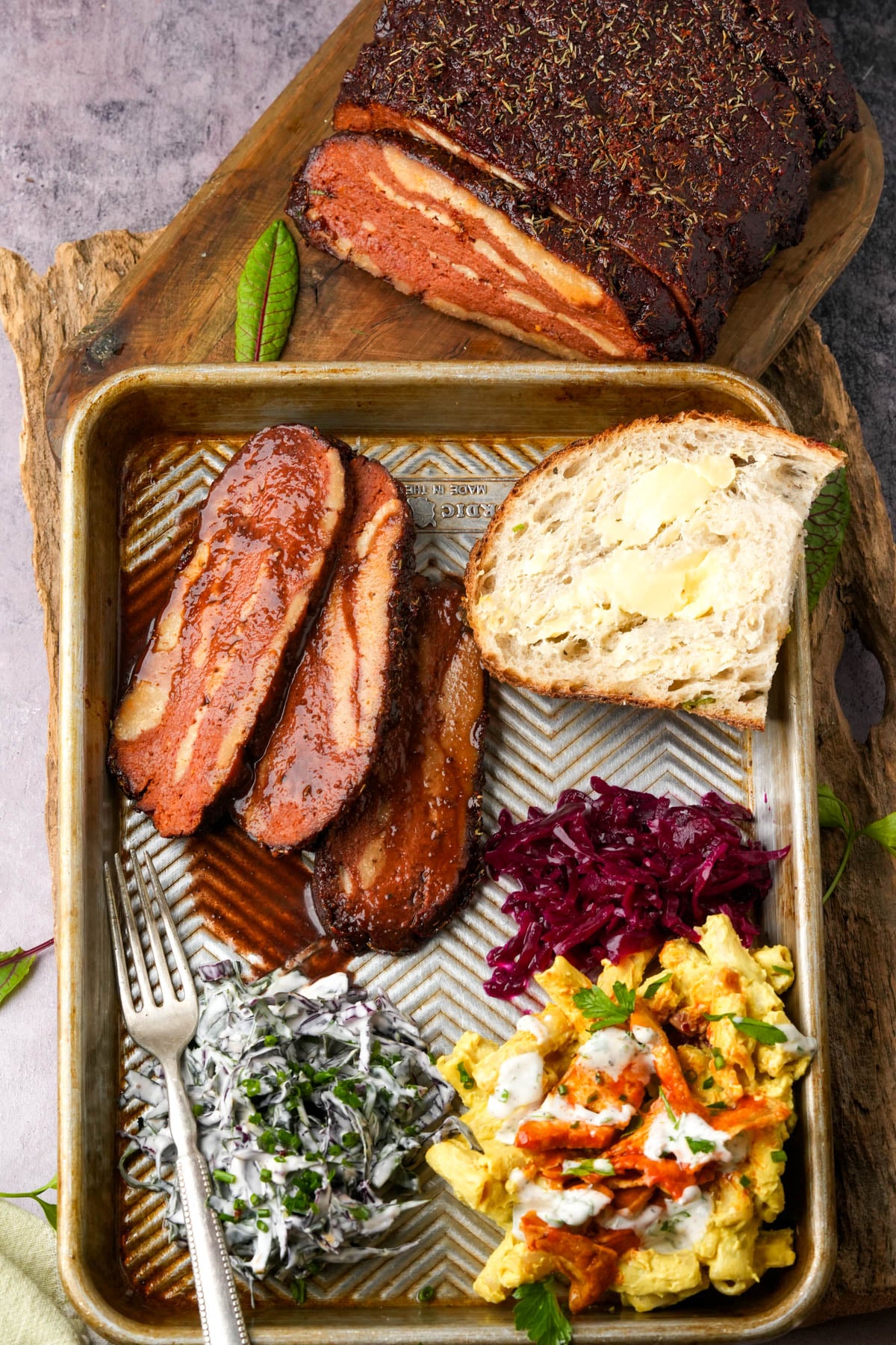 A metal tray holding slices of glazed vegan bbq brisket, a buttered slice of bread, braised red cabbage, a creamy vegetable salad, and a fork, with a large piece of smoked meat on a wooden board above.