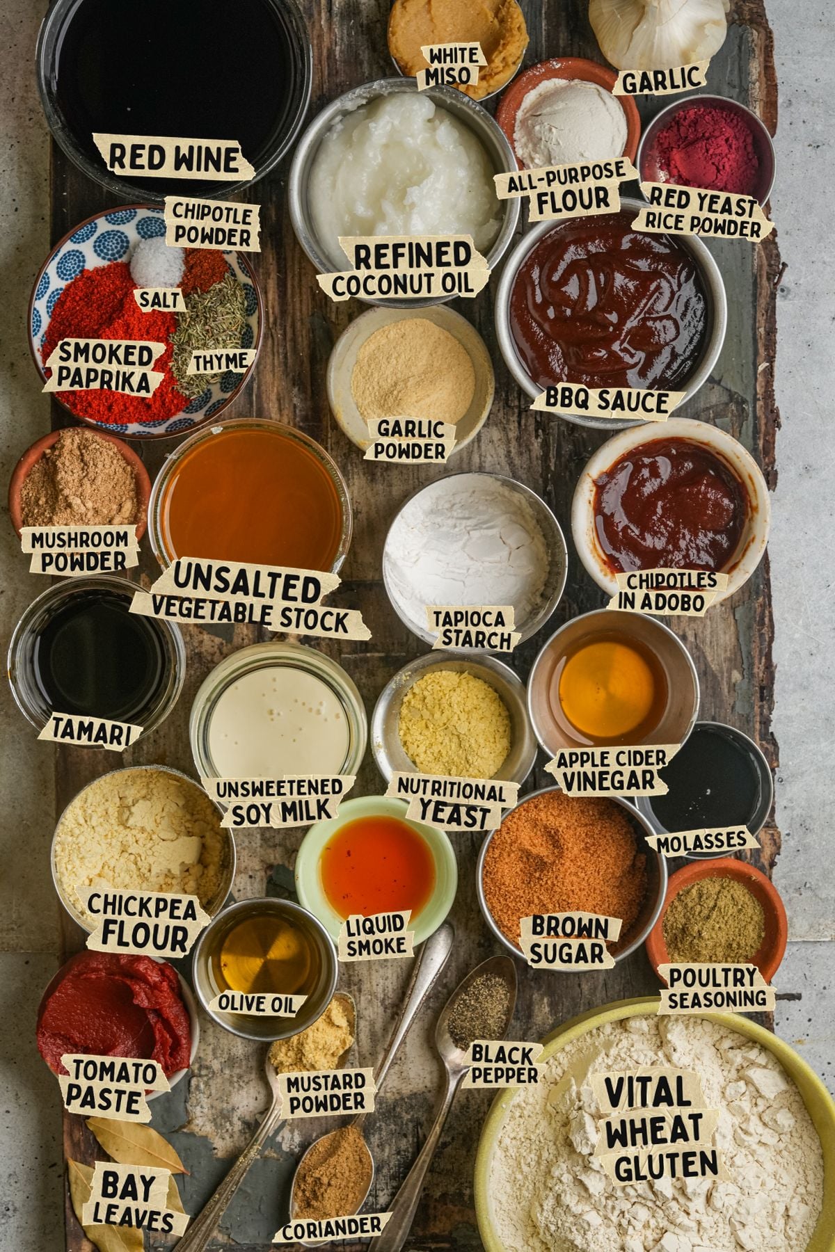 Various vegan bbq brisket ingredients in small bowls and piles on a wooden board, each labeled with its name, including soy milk, tomato paste, miso, spices, and flours.