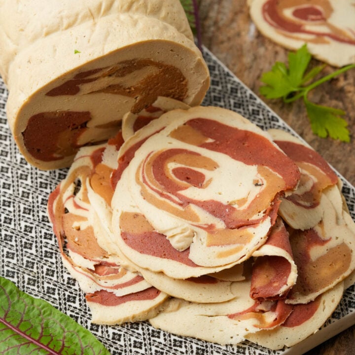 A loaf of plant-based pancetta meat with a red and beige marbled pattern, with several slices arranged in a pile on a patterned tray.
