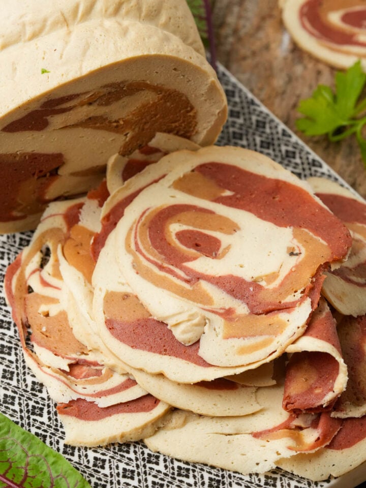 A loaf of plant-based pancetta meat with a red and beige marbled pattern, with several slices arranged in a pile on a patterned tray.