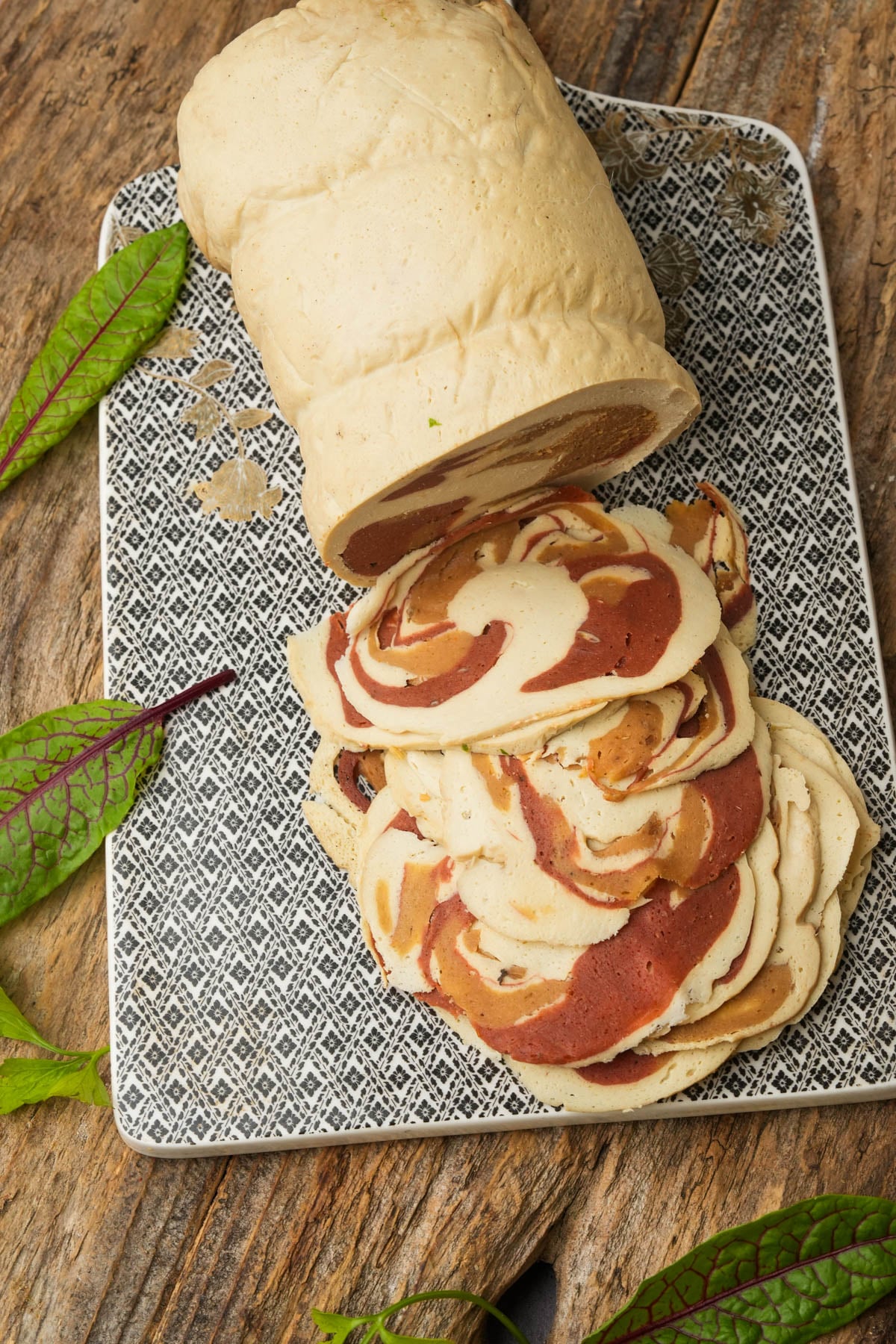A loaf of sliced, swirled vegan pancetta meat with light and dark portions on a patterned serving board.