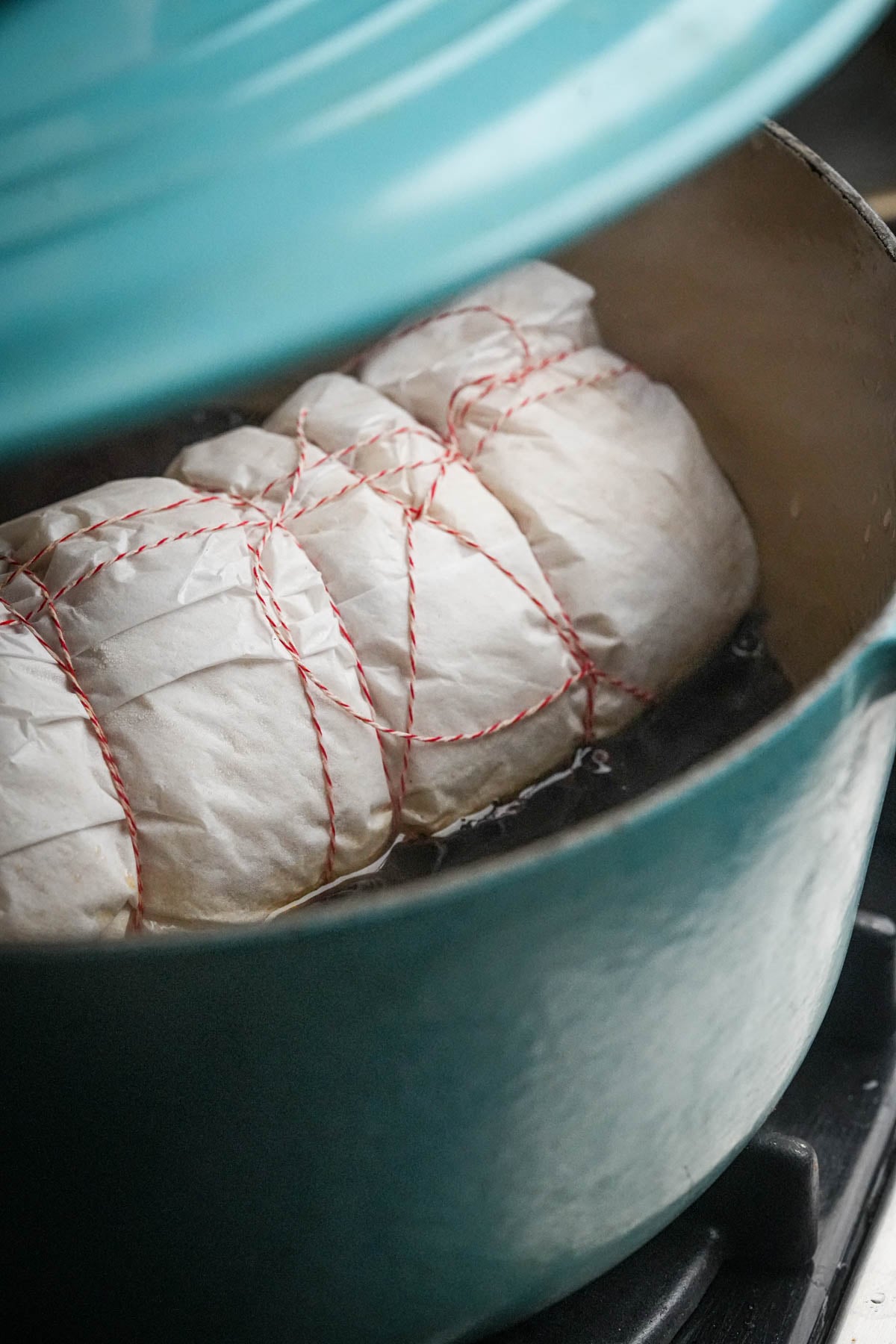 A teal Dutch oven containing raw vegan pancetta dough wrapped in parchment paper and tied with red and white kitchen twine.