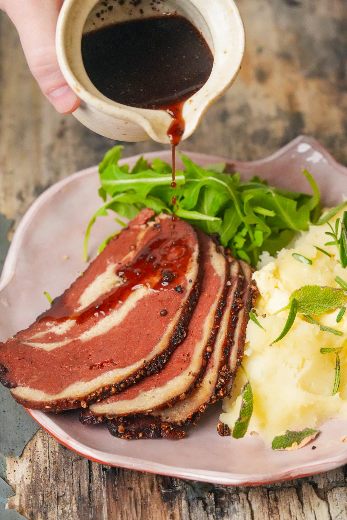 Hand pouring dark sauce over sliced roast meat, served with mashed potatoes and arugula on a plate.