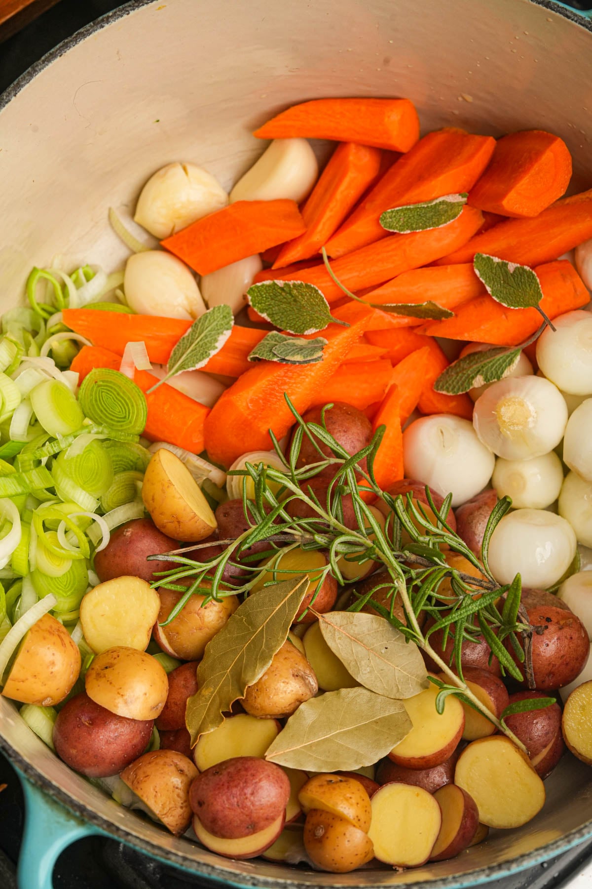Colorful arrangement of chopped carrots, potatoes, pearl onions, leeks, garlic cloves, rosemary, sage, and bay leaves in a large pot, ready for cooking.