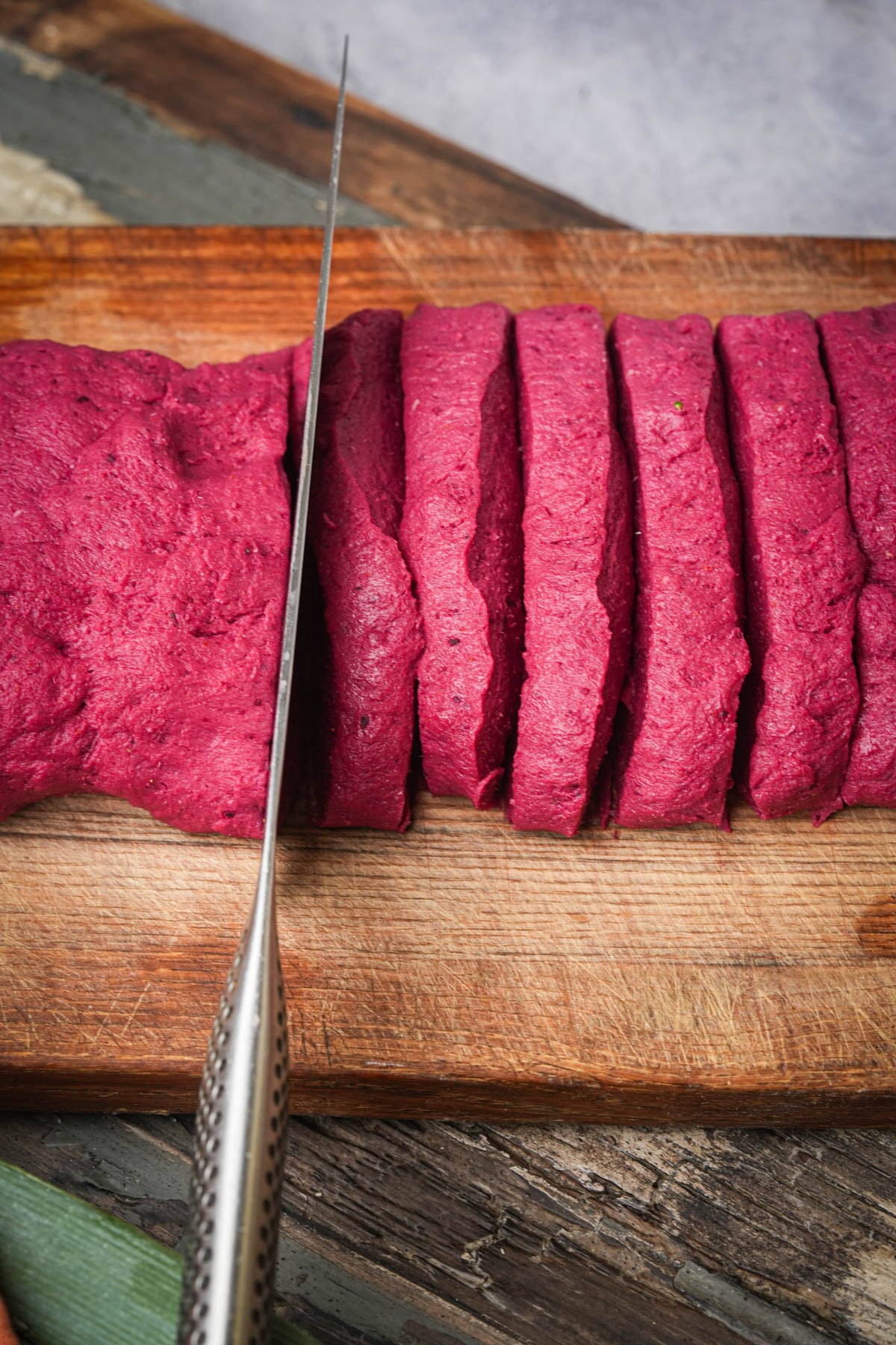 A knife slicing a loaf of bright pink seitan dough into even pieces on a wooden cutting board.