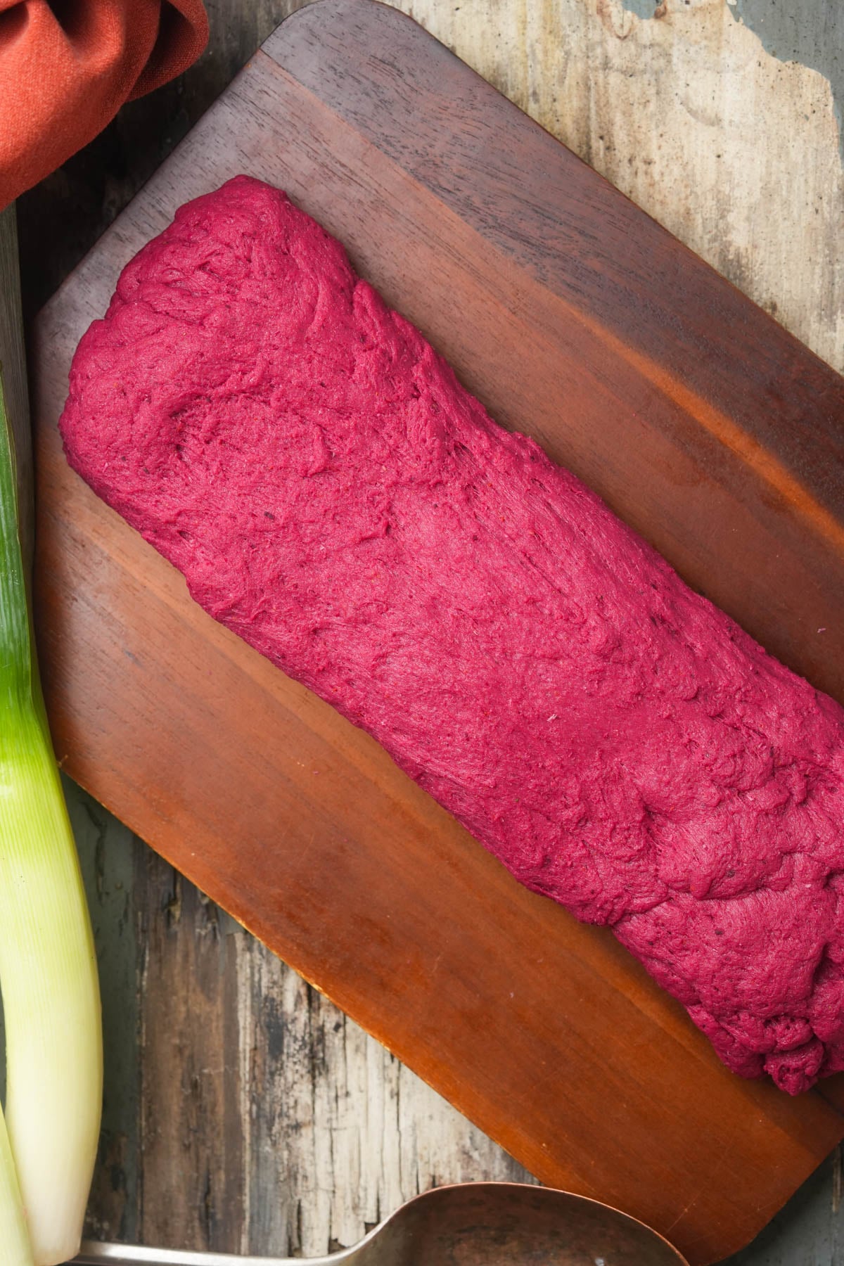 A rectangular log of magenta vegan pot roast setian dough sits on a dark wooden cutting board, next to a leek and a large metal spoon, with a rustic table surface underneath.