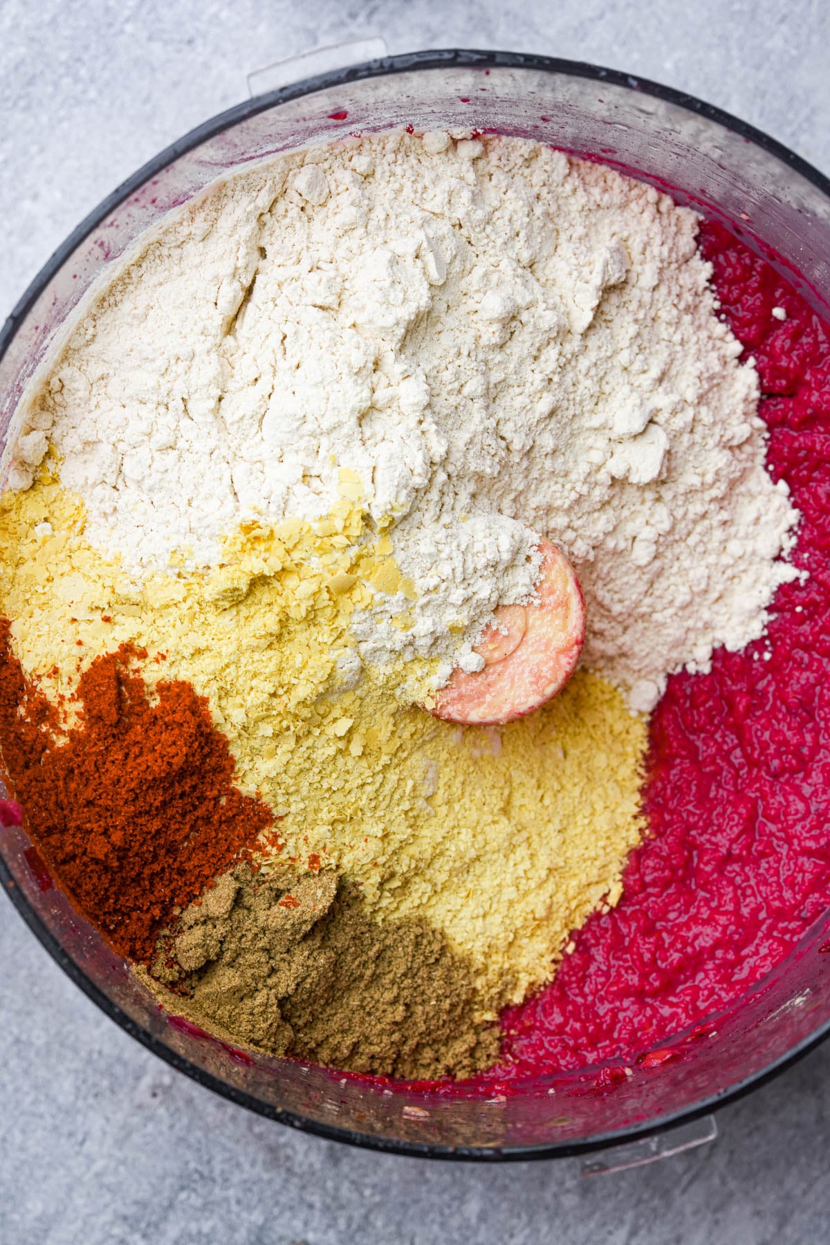 Overhead view of a food processor with vital wheat gluten, nutritional yeast, coriander, paprika, and a bright pink beet puree inside, before blending.