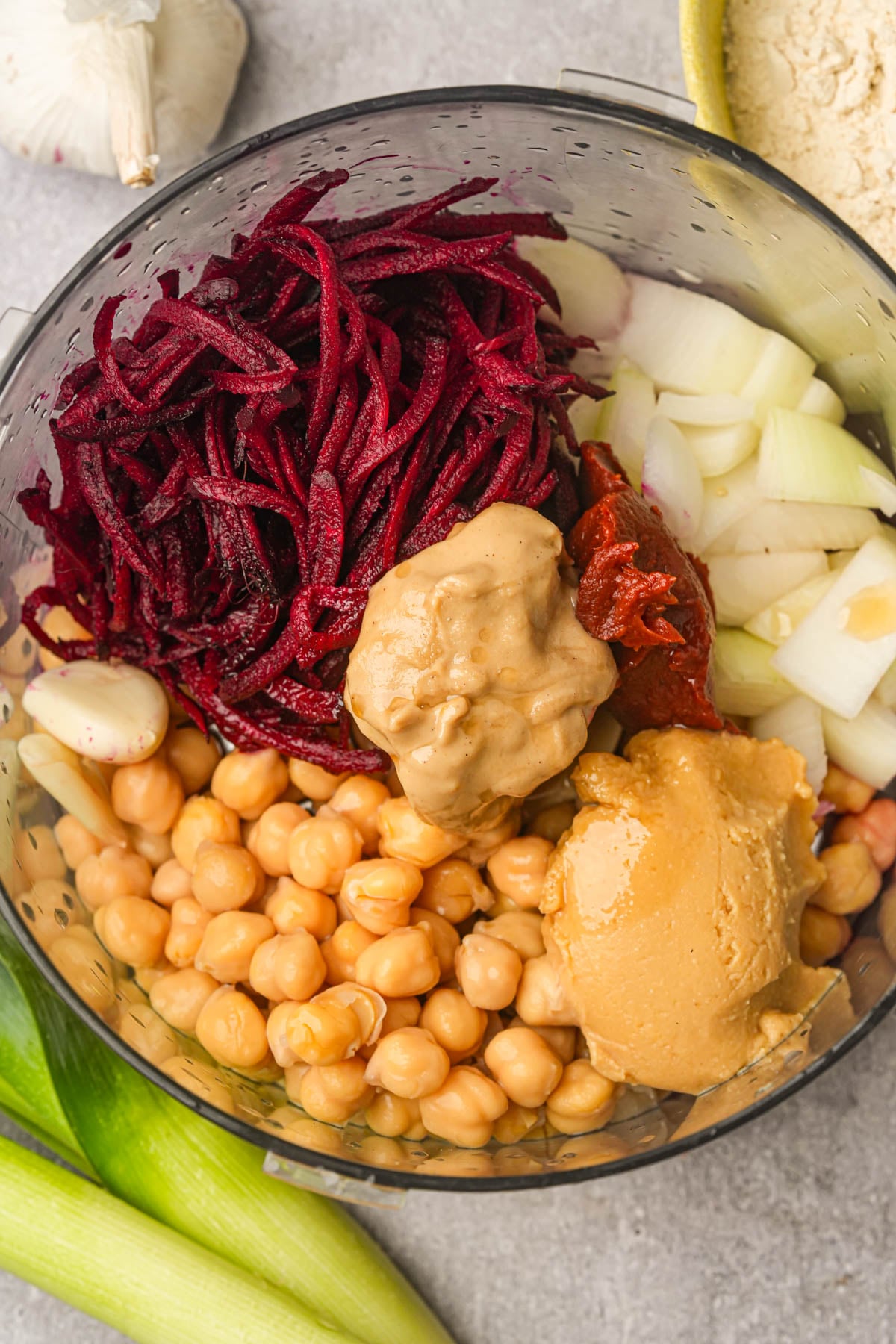 A food processor bowl filled with chickpeas, shredded beets, onions, whole garlic cloves, tahini and tomato paste, ready to be blended.
