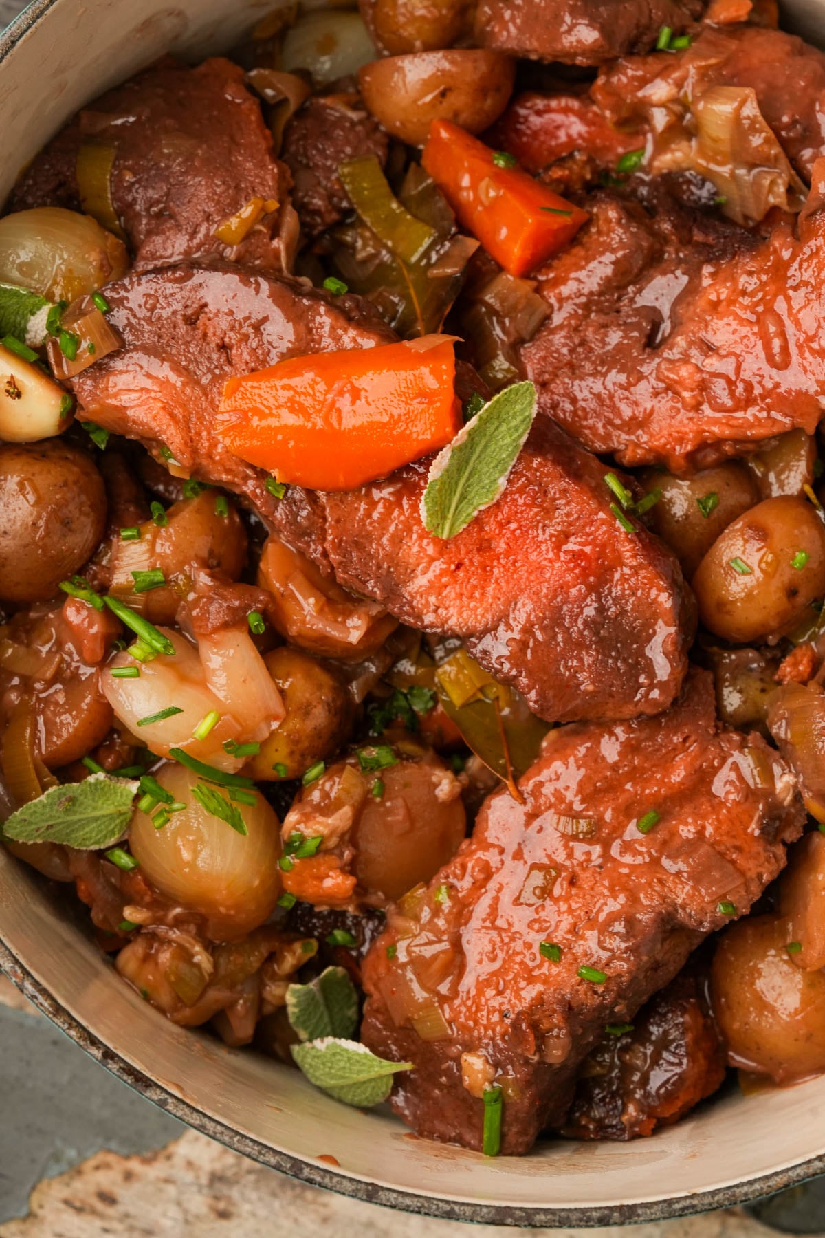 A vegan pot roast with chunks of beef, small potatoes, pearl onions, carrots, and herbs in rich brown sauce, garnished with fresh green herbs.