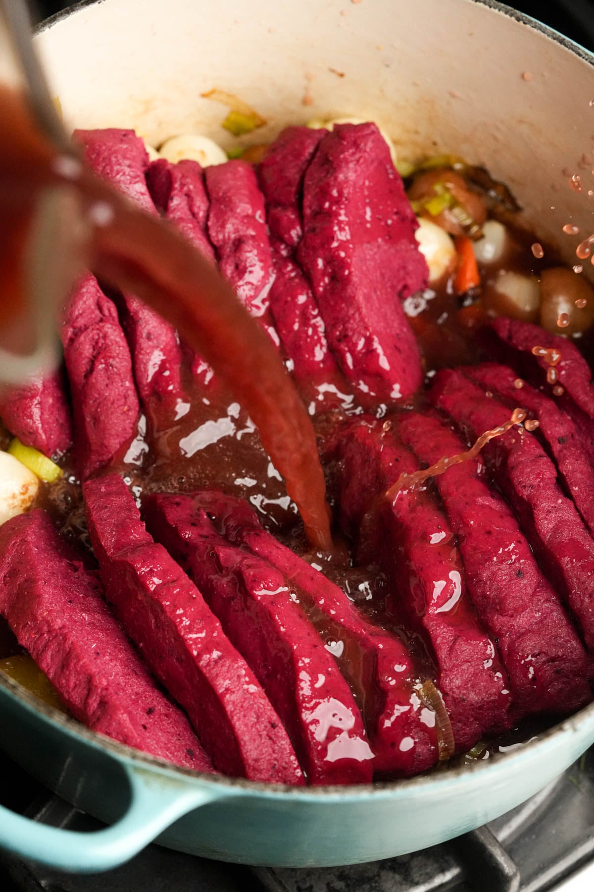 Sliced raw seitan with a deep red color arranged in a pot with vegetables, as a dark liquid, likely broth or wine, is being poured over the vegan pot roast.