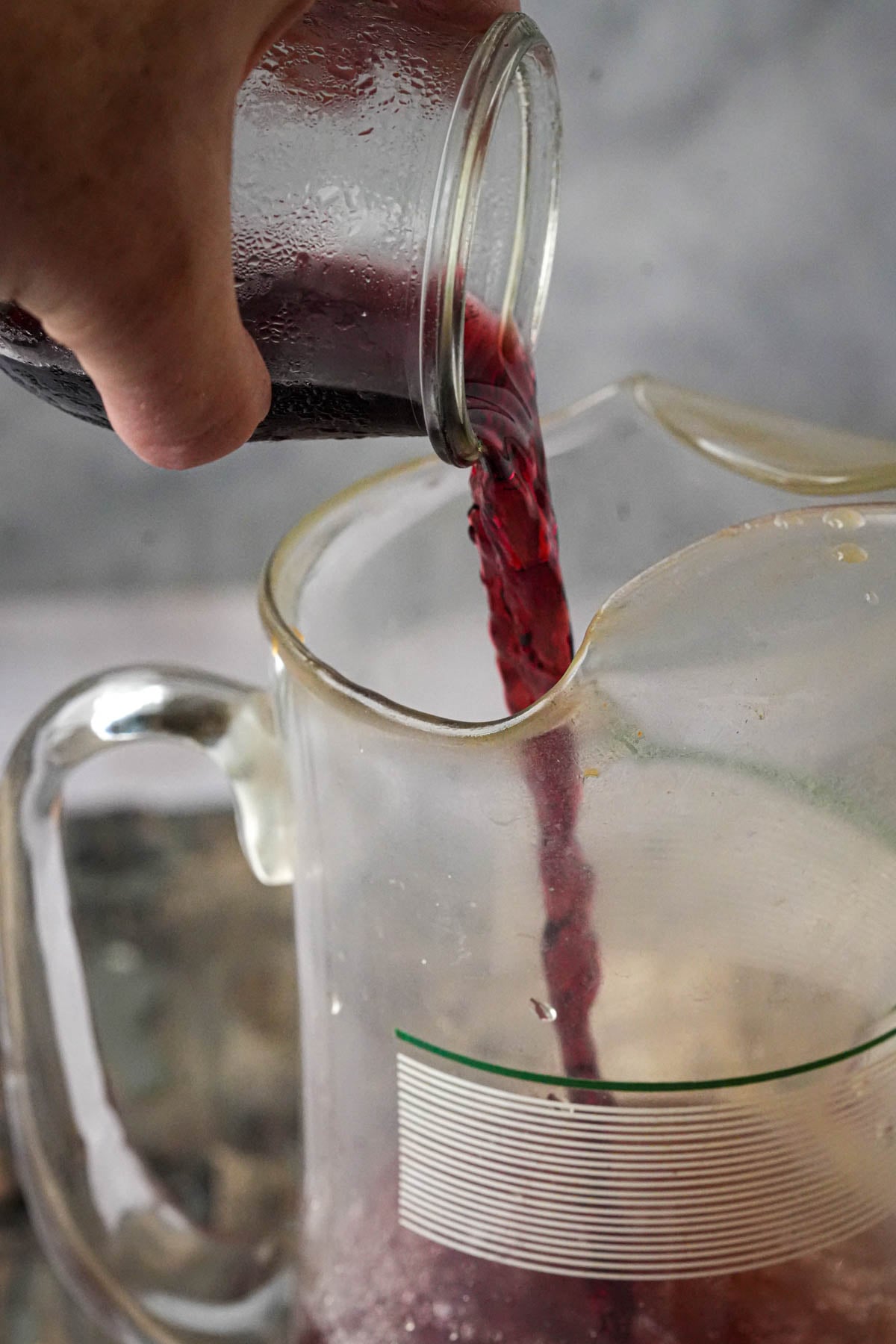 A hand pours red wine from a small glass jar into a clear pitcher with a green-striped label.