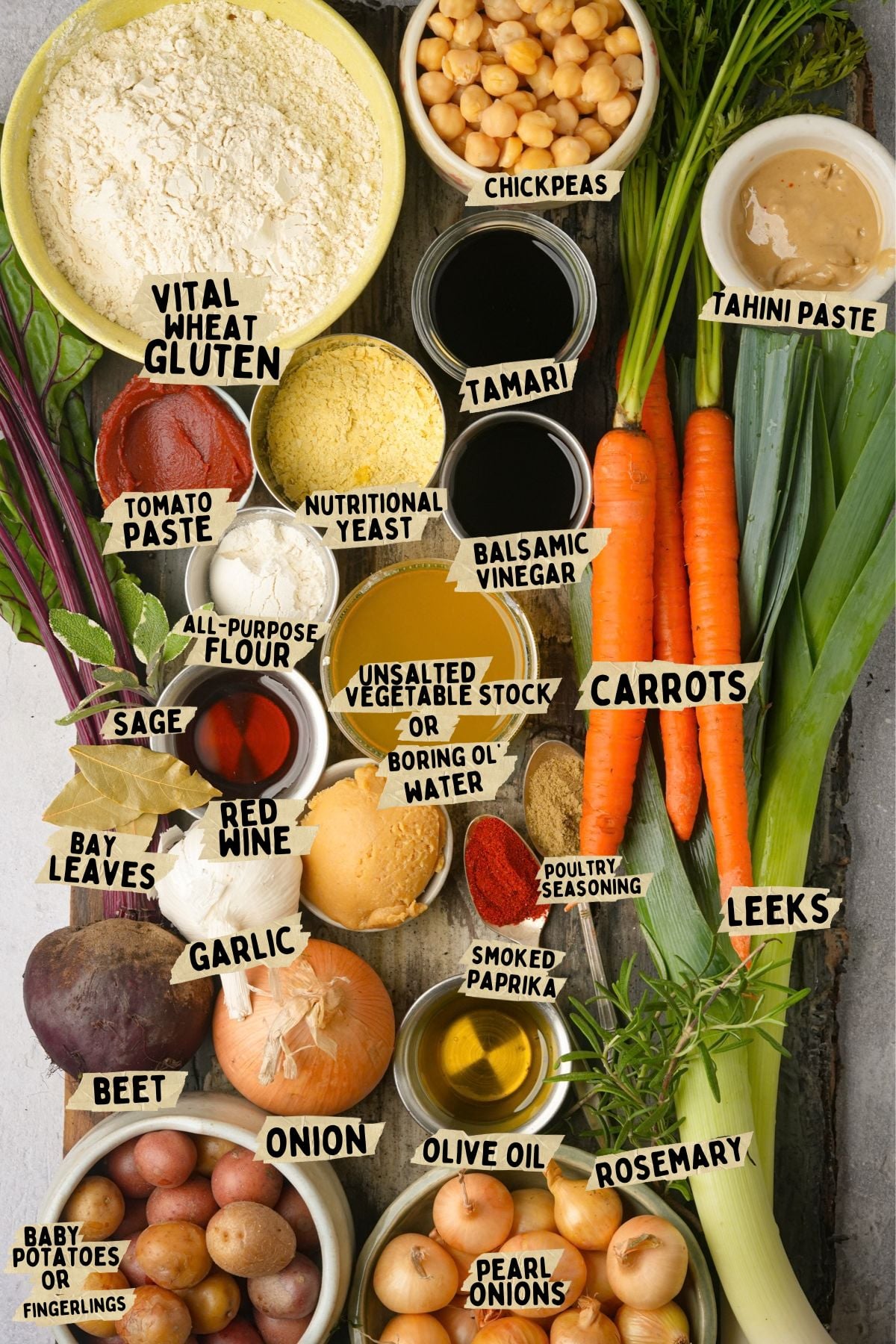 Assorted vegetables, herbs, and ingredients for a vegan pot roast recipe including carrots, leeks, garlic, chickpeas, bay leaves, tahini, olive oil, and seasonings arranged on a table, each labeled with handwritten text naming the item.