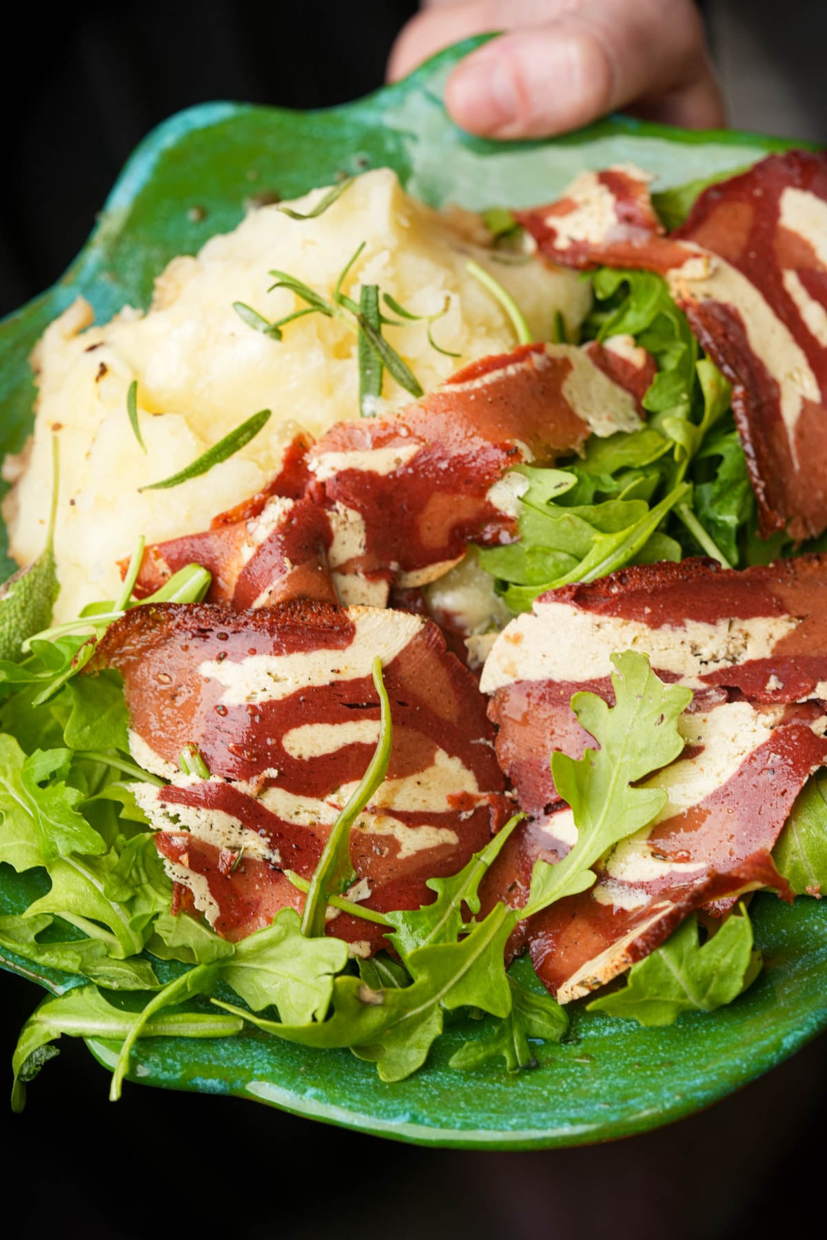 A serving of mashed potatoes topped with marbled cured meat and fresh arugula on a green ceramic plate.