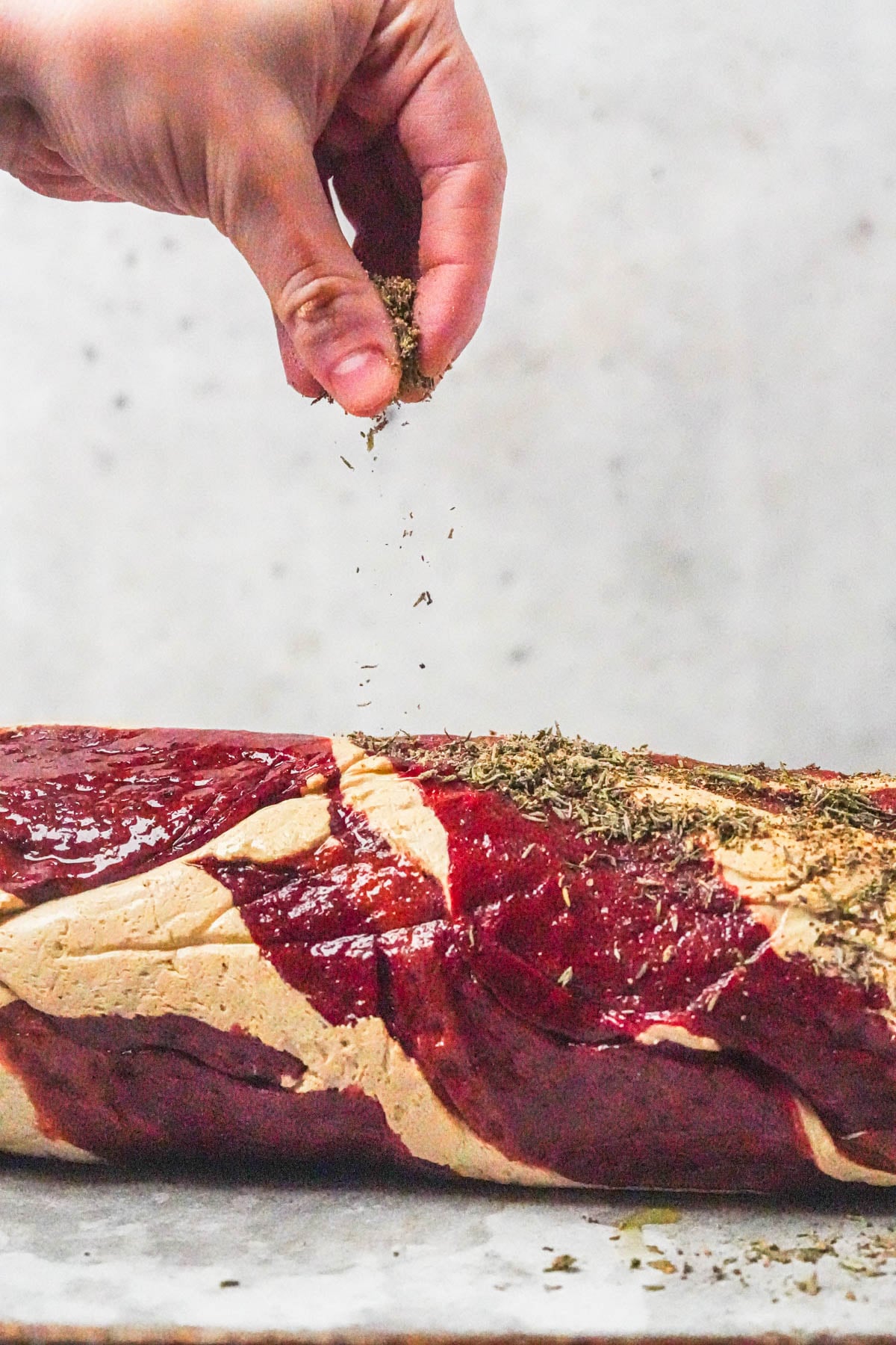 A hand sprinkles dried herbs onto a raw beef tenderloin.
